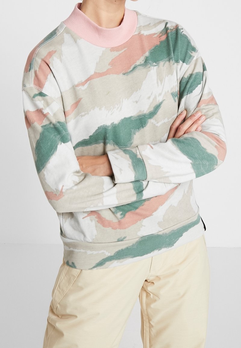 Personne portant un sweat-shirt à col rond à manches longues avec des motifs abstraits verts, beiges et roses et un pantalon couleur crème, bras croisés.