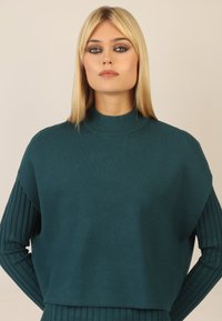 Türkis gestrickter Pullover mit hohem Kragen, der über lange, gerippte Ärmel und eine lockere Passform verfügt. Glatte Textur mit gleichmäßigen Nähten.