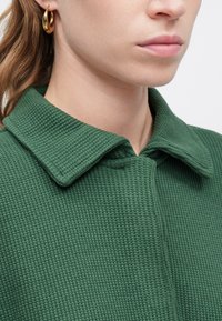 Prenda de color verde con textura y cuello, con cierre de botones, que presenta un patrón de tejido en forma de gofre y un escote redondeado. Pendiente de aro dorado visible.