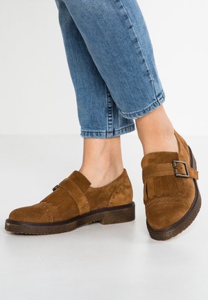 Des pieds chaussés de mocassins en daim marron avec boucle et franges, associés à un jean bleu court sur fond blanc.