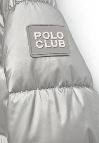 Silverfärgad pufferjacka med en glänsande, slät yta. Har en grå rektangulär etikett med "POLO CLUB" i vit präglad text.