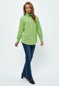 Camicia verde a maniche lunghe con colletto a bottoni, abbinata a jeans a zampa d'elefante blu scuro e stivaletti neri alla caviglia. Tessuto morbido, veste in modo casual.