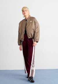 Nabrežna rjava bombaška jakna z zadrgami, v kombinaciji z maroon trenirko, ki ima bele stranske črte in logotip Adidas. Črne superge z gumijastim podplatom.