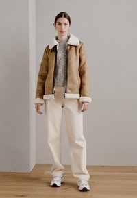 Giacca di shearling marrone con colletto e polsini bianchi, abbinata a pantaloni crema e sneakers bianche. Lo sfondo è una parete grigia uniforme.