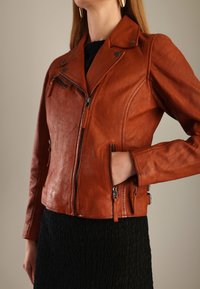 Kvinde iført en rustfarvet læderjakke med lynlåslommer over en sort struktureret kjole, hånd i jakken lommen, mod en neutral baggrund.