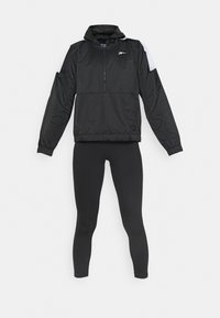 Coupe-vent noir à capuche avec fermeture éclair sur la moitié et détail blanc aux épaules, associé à un legging noir ajusté sur fond uni.