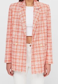 Donna che indossa un blazer a quadri arancione e bianco sopra un crop top bianco e pantaloni bianchi a vita alta su uno sfondo neutro.