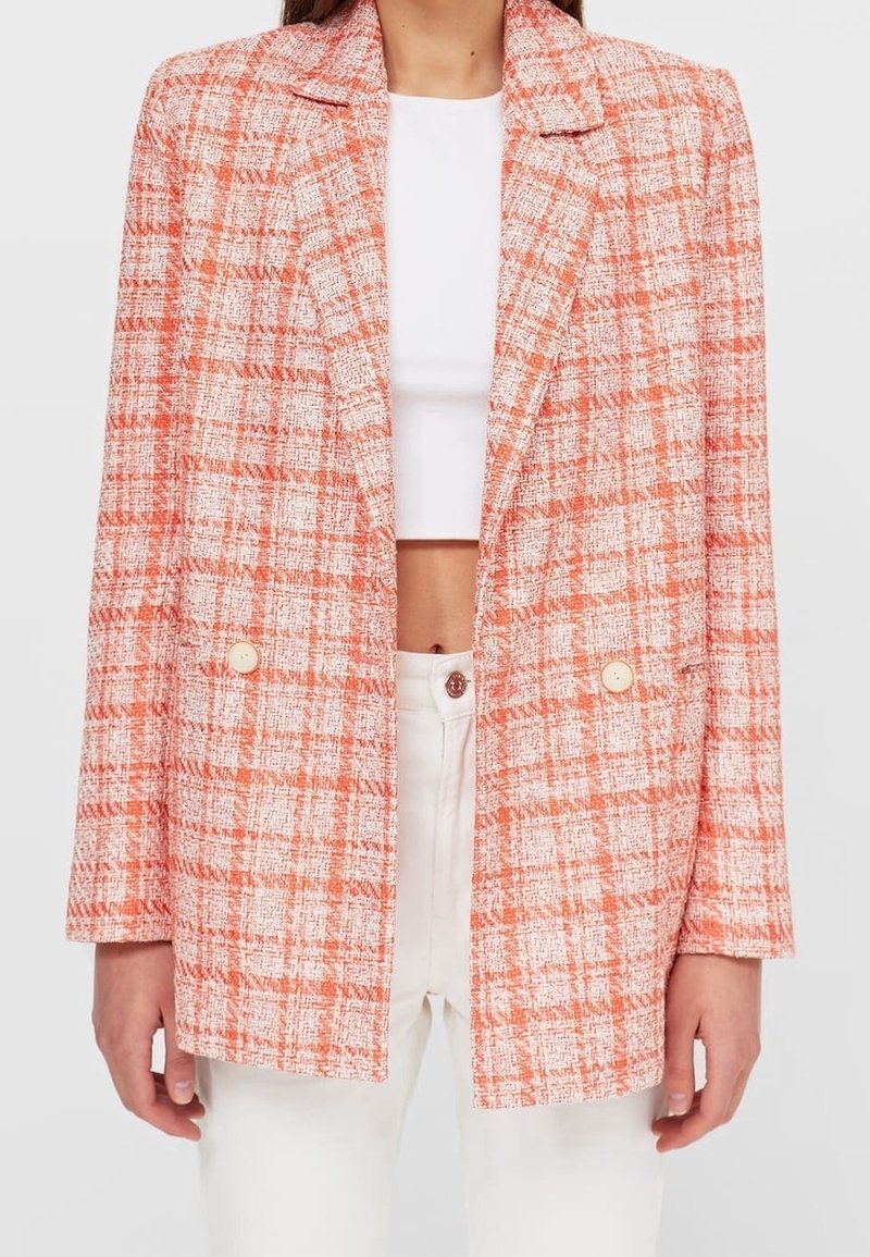 Donna che indossa un blazer a quadri arancione e bianco sopra un crop top bianco e pantaloni bianchi a vita alta su uno sfondo neutro.