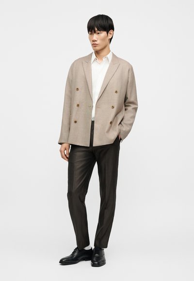 Μπεζ σακάκι διπλής σειράς με καφέ κουμπιά, συνδυασμένο με λευκό πουκάμισο και σκουρόχρωμο παντελόνι με σχέδια. Μαύρα παπούτσια με κορδόνια ολοκληρώνουν την εμφάνιση.