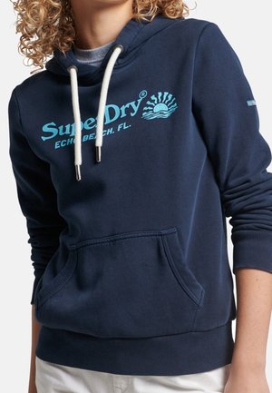 Personne aux cheveux bouclés portant un sweat à capuche SuperDry bleu marine avec des cordons blancs et une poche avant, associé à un pantalon blanc.