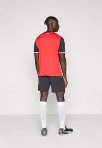 Κόκκινη φανέλα ποδοσφαίρου με μαύρα μανίκια, λευκές λεπτομέρειες στον γιακά και τις μανσέτες. Μαύρο σορτς, λευκές κάλτσες και μαύρα ποδοσφαιρικά παπούτσια.