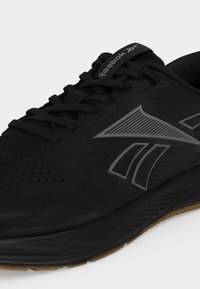 Μαύρο αθλητικό παπούτσι Reebok με γκρι λογότυπο, μαύρα κορδόνια και σόλα σε χρώμα καουτσούκ, ορατό από μπροστινή πλευρική γωνία.