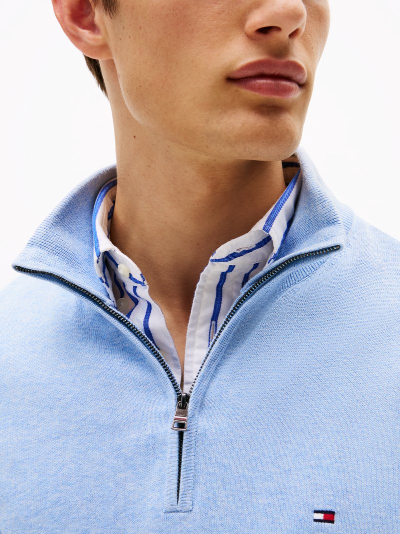 Hellblaues Zip-up Sweatshirt aus weichem Stoff, mit hohem Kragen und einem darunter getragenen weißen Hemd mit blauen Streifen. Kleines Logo auf dem Sweatshirt.