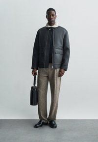 Čierna kožená bunda s ovčia golierom, dve predné vrecká, v kombinácii s textúrovanými hnedými nohavicami a čiernymi topánkami. Nesie čiernu tašku.