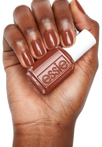 Essie-Nagellack in einem Braunton, der in einer Hand gehalten wird; die Nägel sind im gleichen Farbton lackiert. Glänzende Finish, quadratische Flasche mit einem weißen Deckel.