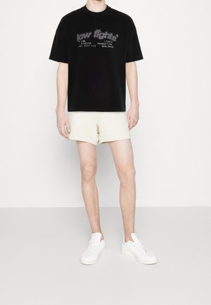 Zwart T-shirt met korte mouwen en geborduurde grafische tekst. Gecombineerd met beige, gestructureerde shorts. Model draagt witte sneakers.