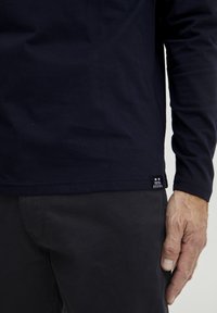 Tengerészkék, hosszú ujjú, pamut ing, egyenes aljjal, kicsi márkás címkével. A modellen sötét nadrág van, a keze az oldalán pihen.