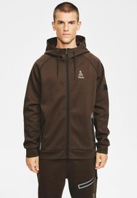 Brązowa bluza z kapturem na suwak wykonana z miękkiego materiału, z kapturem ze sznurkiem. Na piersi znajduje się logo, a przy zamkach błyskawicznych kontrastujące akcenty.