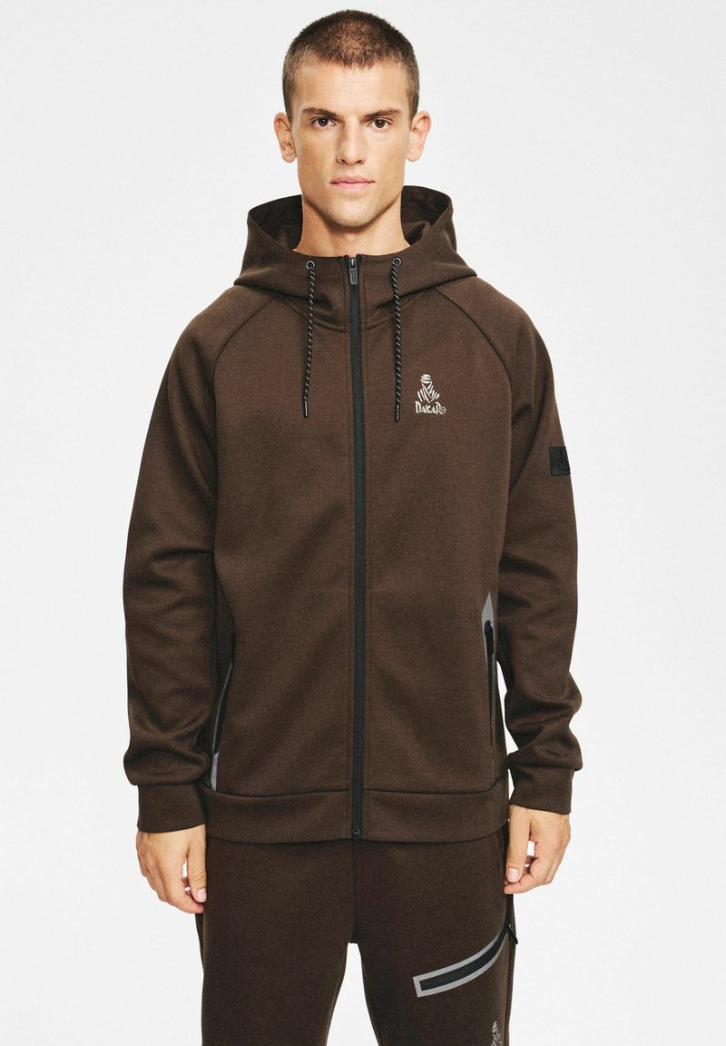 Brązowa bluza z kapturem na suwak wykonana z miękkiego materiału, z kapturem ze sznurkiem. Na piersi znajduje się logo, a przy zamkach błyskawicznych kontrastujące akcenty.