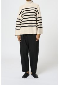 Beżowy golf z czarnymi poziomymi paskami, o oversize'owym kroju i szerokich rękawach, zestawiony z czarnymi luźnymi spodniami oraz czarnymi wsuwanymi butami.