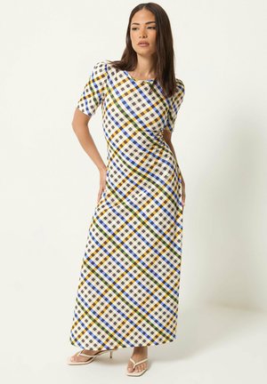 Femme portant une robe longue à carreaux multicolores avec manches courtes et sandales à talons à bout carré, posant devant un fond clair uni.