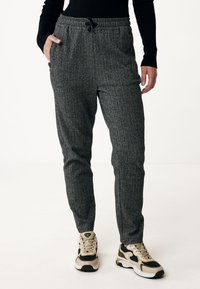 Šedé jogger kalhoty s herringbone vzorem, elastickým pasem a šňůrkou na stažení. Boční kapsy a zúžené nohavice. Na sobě má béžové a černé tenisky.