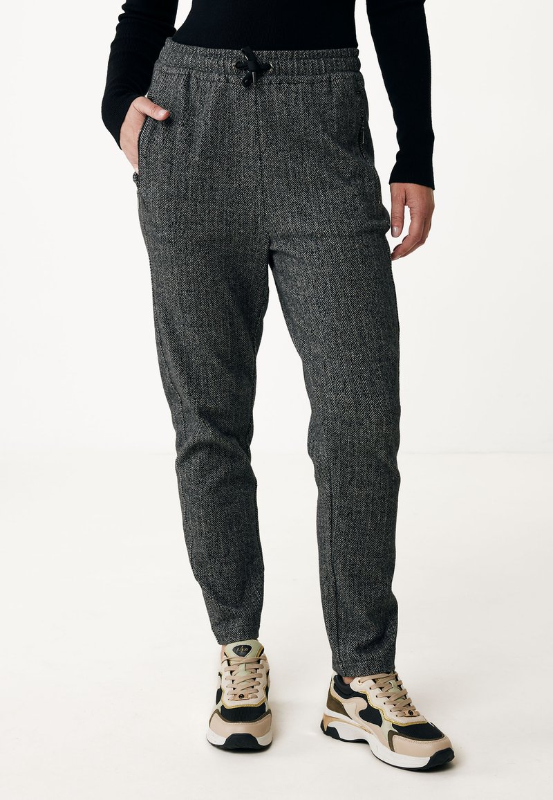 Šedé jogger kalhoty s herringbone vzorem, elastickým pasem a šňůrkou na stažení. Boční kapsy a zúžené nohavice. Na sobě má béžové a černé tenisky.