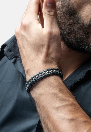 Braided black leather bracelet with silver metal accents, featuring a smooth texture and a layered design, worn on a wrist.