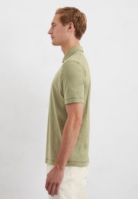 Jasnozielona koszulka polo z klasycznym kołnierzykiem i krótkimi rękawami, wykonana z miękkiego, strukturalnego materiału, prezentowana na modelu mężczyźnie w profilu.