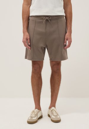 Man met taupekleurige shorts met trekkoord en witte sneakers, staand tegen een effen achtergrond.