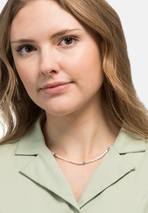 Camicia verde chiaro con colletto, indossata con una collana di perle con accenti in argento. Texture del tessuto liscia, primo piano della zona del collo.