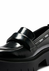 Mocassin en cuir noir brillant avec une semelle épaisse à crampons et un détail de la marque "HUGO" sur le côté près de l'ouverture.