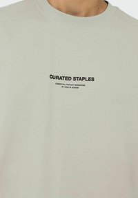 Lichtgroen katoenen T-shirt met rond de hals, voorzien van zwarte tekst die "CURATED STAPLES" leest en extra branding eronder in een eenvoudig lettertype.