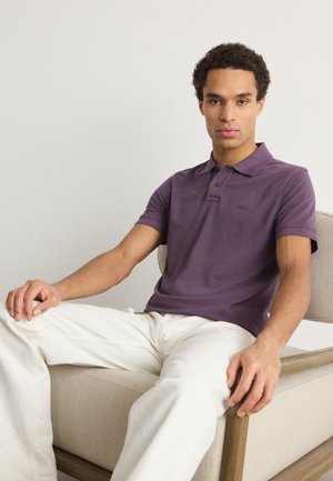 Lila Poloshirt mit Kragen und kurzen Ärmeln, kombiniert mit beigen Hosen. Das Modell sitzt auf einem hellen Stuhl.