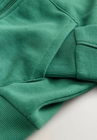 Tissu de sweat-shirt vert avec une texture lisse, des poignets côtelés et un détail de couture plate. Gros plan montrant la matière et les coutures.