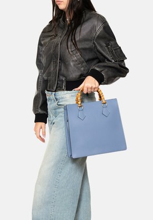 Sac à main en cuir bleu avec une forme structurée, poignées style bambou et accents angulaires, tenu dans une main, à côté d'une veste bomber noire et d'un jean clair.