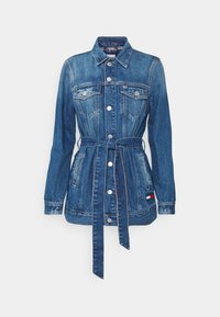 Chaqueta de mezclilla con cintura ajustada, cierre de botones, dos bolsillos en el pecho y un pequeño parche de logo en rojo, blanco y azul en la parte inferior izquierda.