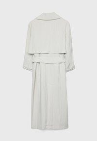 Long trench-coat blanc avec grand col, empiècement arrière, ceinture à la taille et poignets retroussés sur les manches, vu de dos sur fond uni.