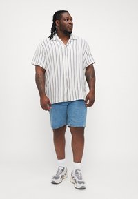 Camisa de manga corta a rayas en tela ligera, con rayas verticales azules y moradas, combinada con shorts de mezclilla. Deportivas grises y calcetines blancos.