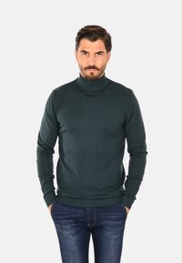 Maglione dolcevita verde scuro in materiale a maglia, design aderente con maniche lunghe, polsini e orlo a coste, indossato con jeans scuri in denim.