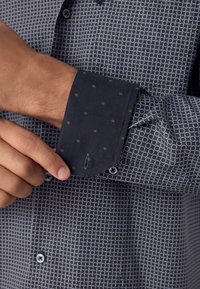 Marineblauwe shirt met een geometrisch patroon in lichtblauw en wit, met een contrasterende marineblauwe manchetten versierd met kleine gouden stippen en knoopdetail.