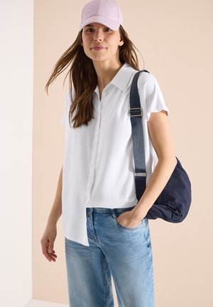 Kvinde iført hvid kortærmet skjorte med knapper, lyserød kasket, blå jeans, bærende på en marineblå skuldertaske med bred rem, stående mod en beige baggrund.