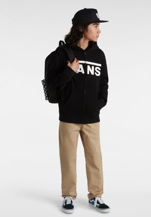 Czarna bluza z zamkiem błyskawicznym z dużym białym logo "VANS", beżowe spodnie, czarny plecak w kratę oraz sneakersy z niebieskimi akcentami.