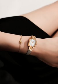 Montre ton or avec un cadran carré, un fond blanc et des éléments en or. Dotée d'un bracelet gris clair. Accompagnée d'un délicat bracelet en or.