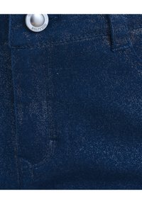 Gros plan sur un tissu en denim bleu foncé avec un bouton en métal argenté portant l'inscription « GULLIVER » et des coutures détaillées près d'une boucle de ceinture.