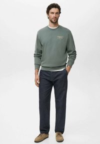 Light green sweatshirt with ribbed cuffs and neckline, layered over a white shirt, paired with dark blue trousers and beige shoes.