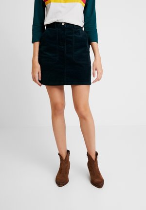 Vrouw draagt een donkere corduroy minirok met voorzakken, een colorblock top met groene mouwen, en bruine suède enkellaarsjes.