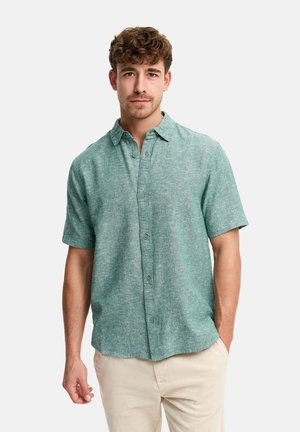 Homme aux cheveux bouclés portant une chemise verte à manches courtes boutonnée et un pantalon beige, debout avec une main dans la poche.