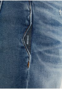 Světle modré džíny z denimu s tónovaným prošitím. Na boku štítek s nápisem "CHASIN". Vybledlé detaily u kapsy; celkově hladká textura.