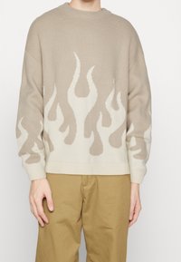 Beżowy sweter dzianinowy z motywami płomieni w jaśniejszym odcieniu. Okrągły dekolt i ściągacze na rękawach, w połączeniu z khaki spodniami.